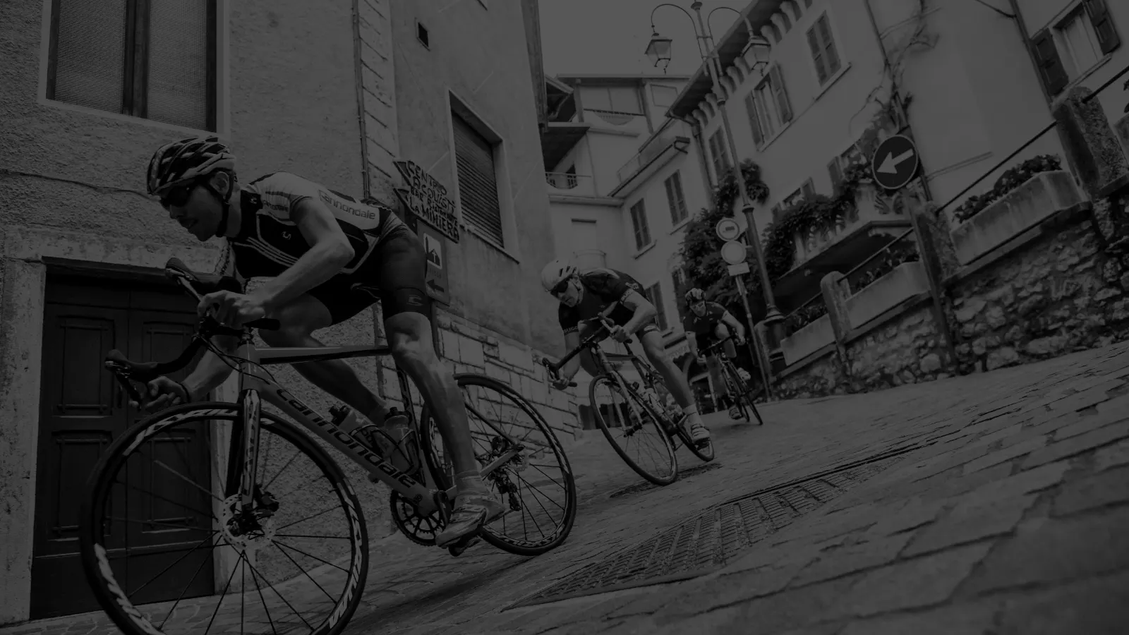 Due ciclisti su strada in discesa tra le case di un paese, in bianco e nero